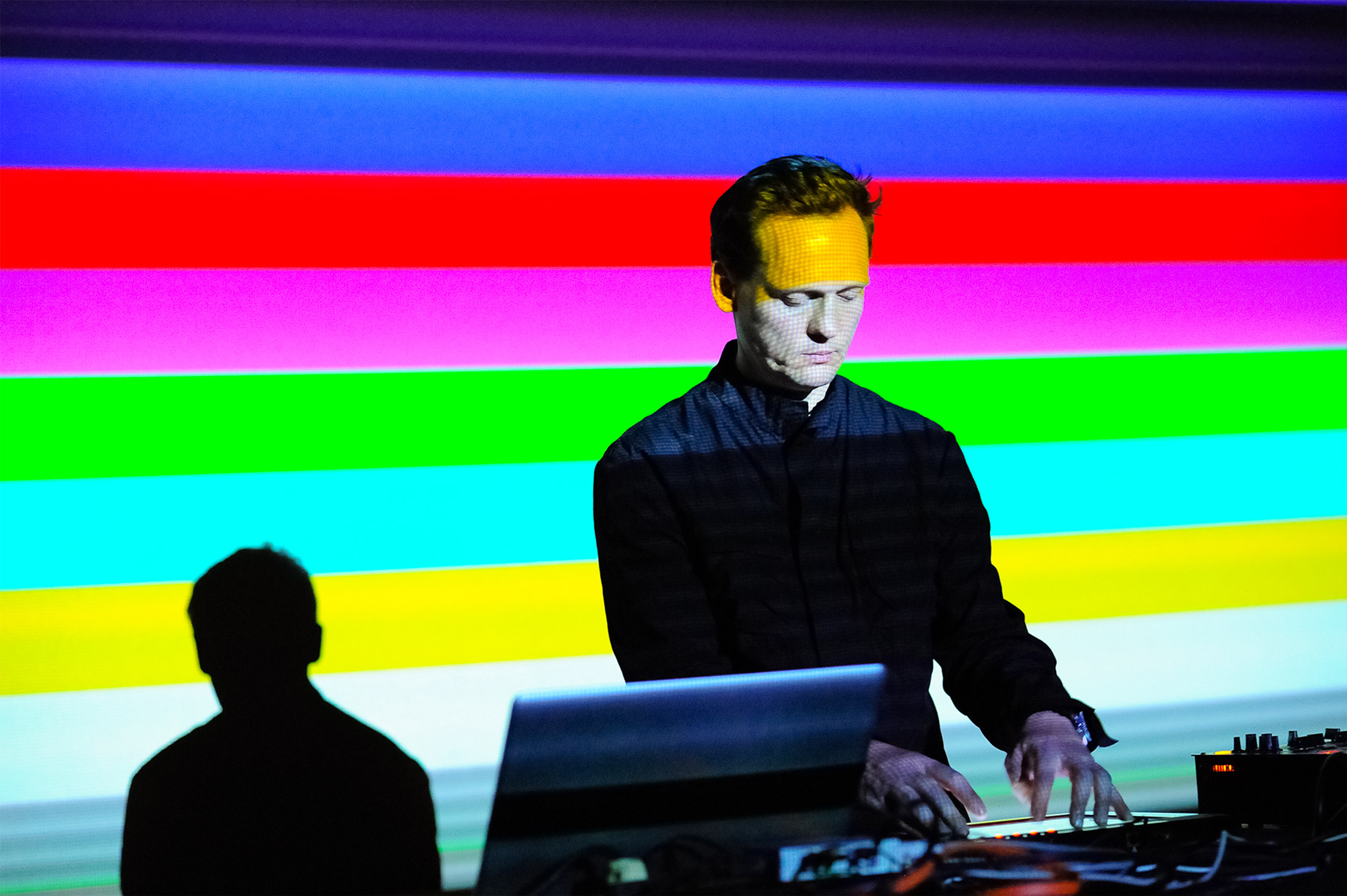 Shows Carsten playing music from a laptop in the middle, wearing a black shirt. Then the background is a bright blue, red, green, light green and yellow stripped background, like a TV error screen.