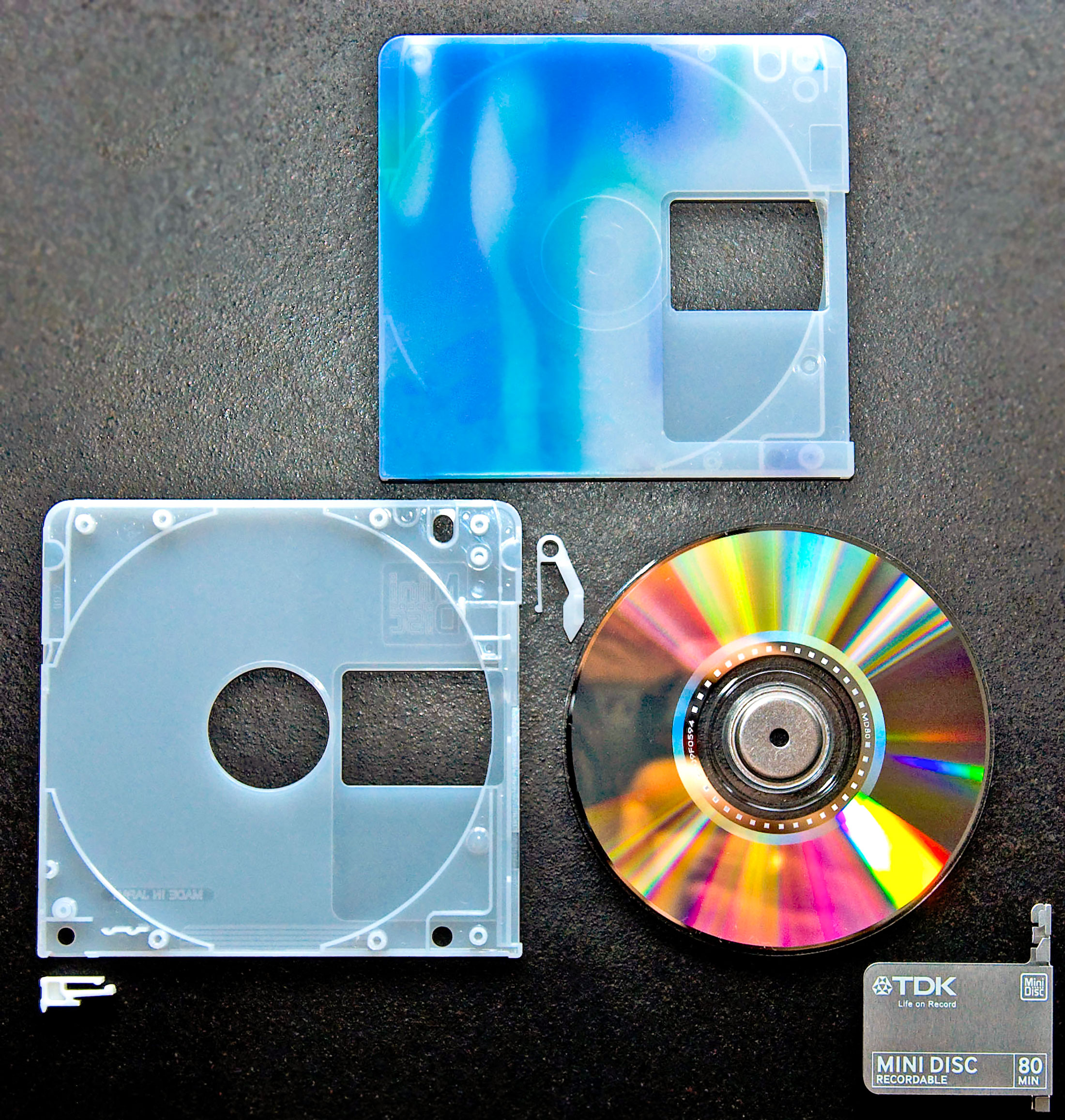 Photo of a minidisc that has been deconstructed. On the top shows the blue plastic outside covering. On the bottom left shows the inside white plastic container. To the bottom right, shows the actual round minidisc. Then below this is the metal slider, that protects the reading and writing of the mindisc.