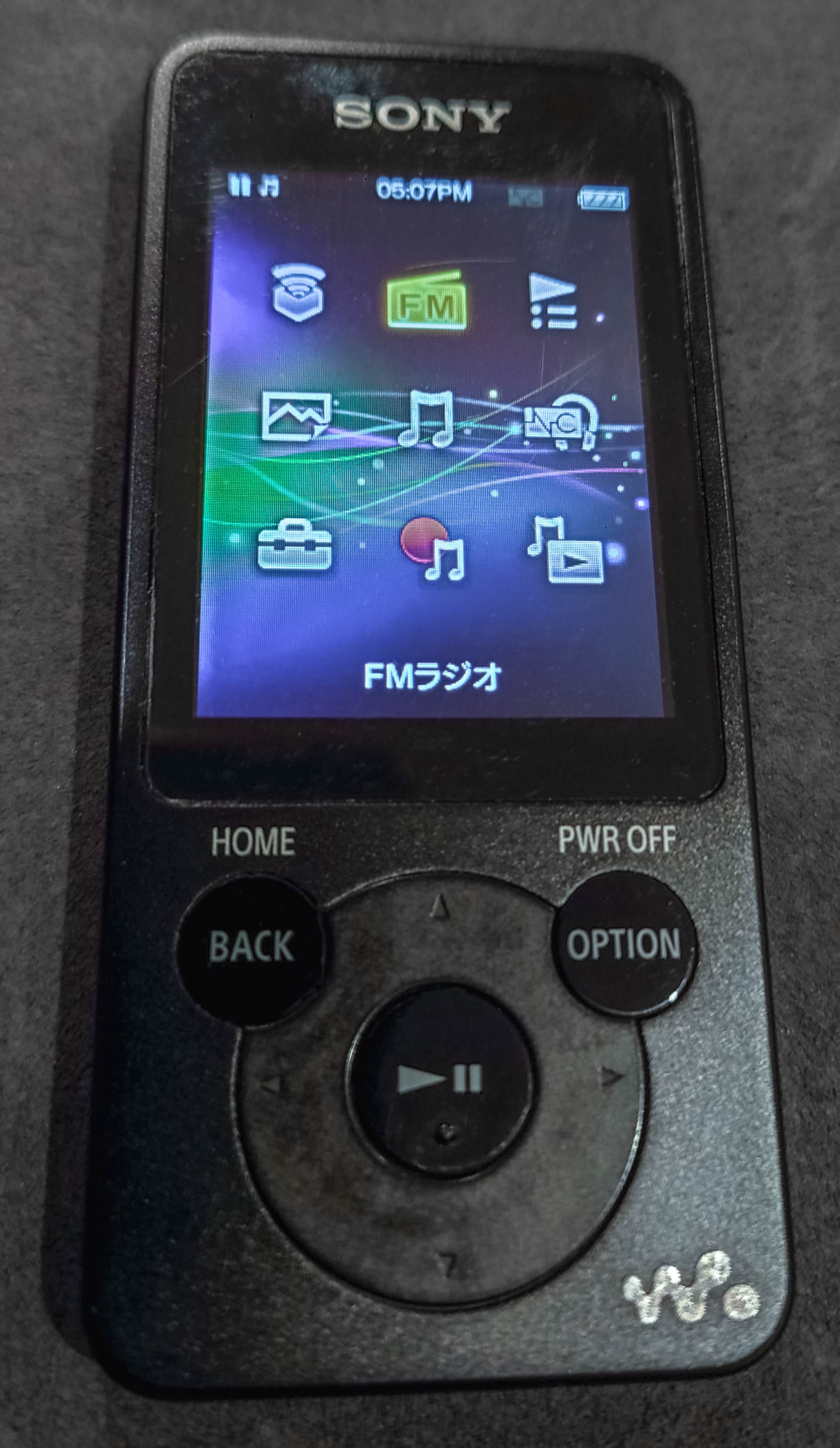 Front on up-close photograph of the black digital Walkman. Show a digital screen covering the top half of the Walkman, it has various options on it. Then below that, a back, option and home control interface.