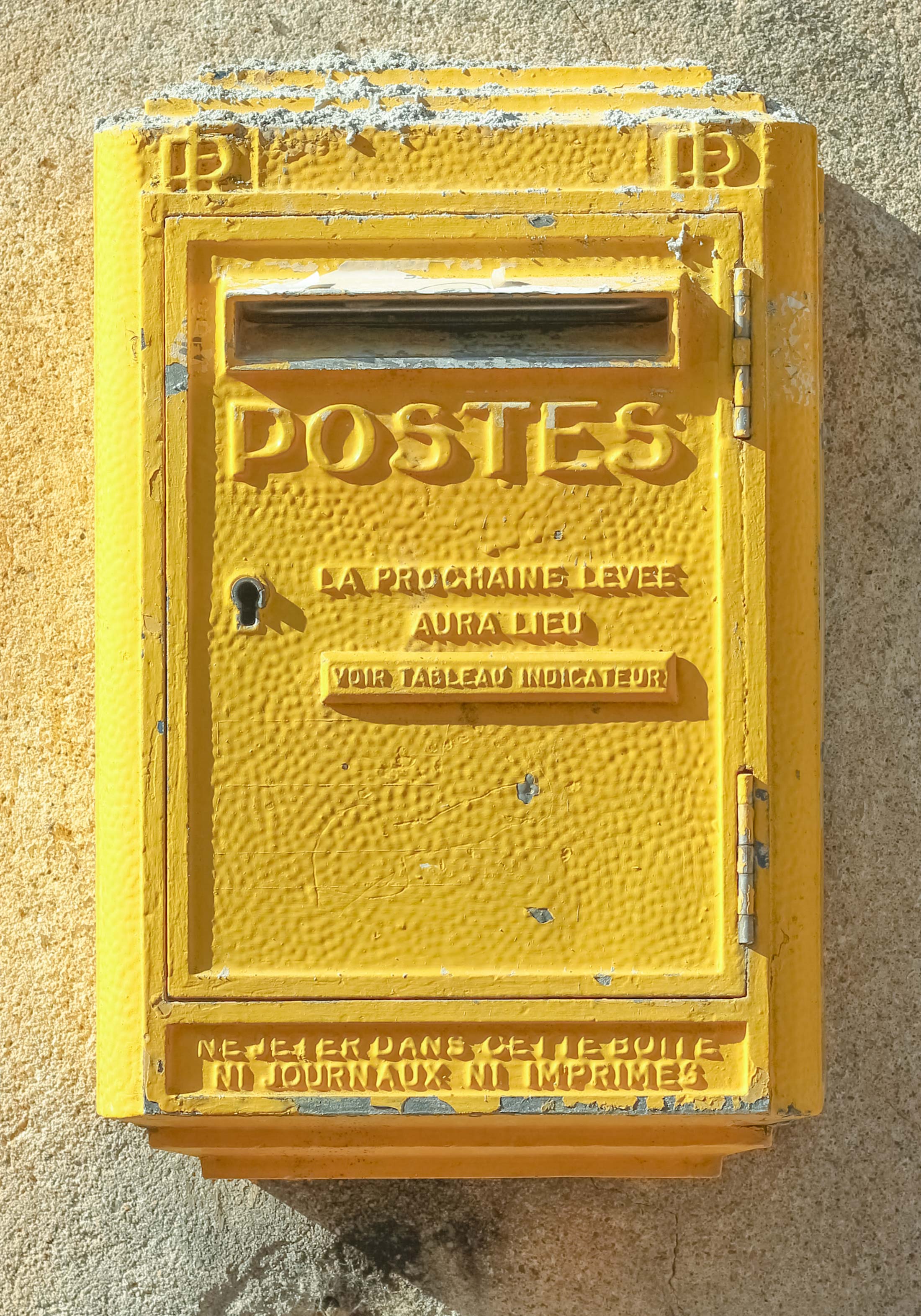 Bright yellow old French steel mailbox, on a wall. Has a small entry for letters at the top, then below in capital letter the word ‘POSTES, LA PROCHAINE LEVEE AURA LIED’.