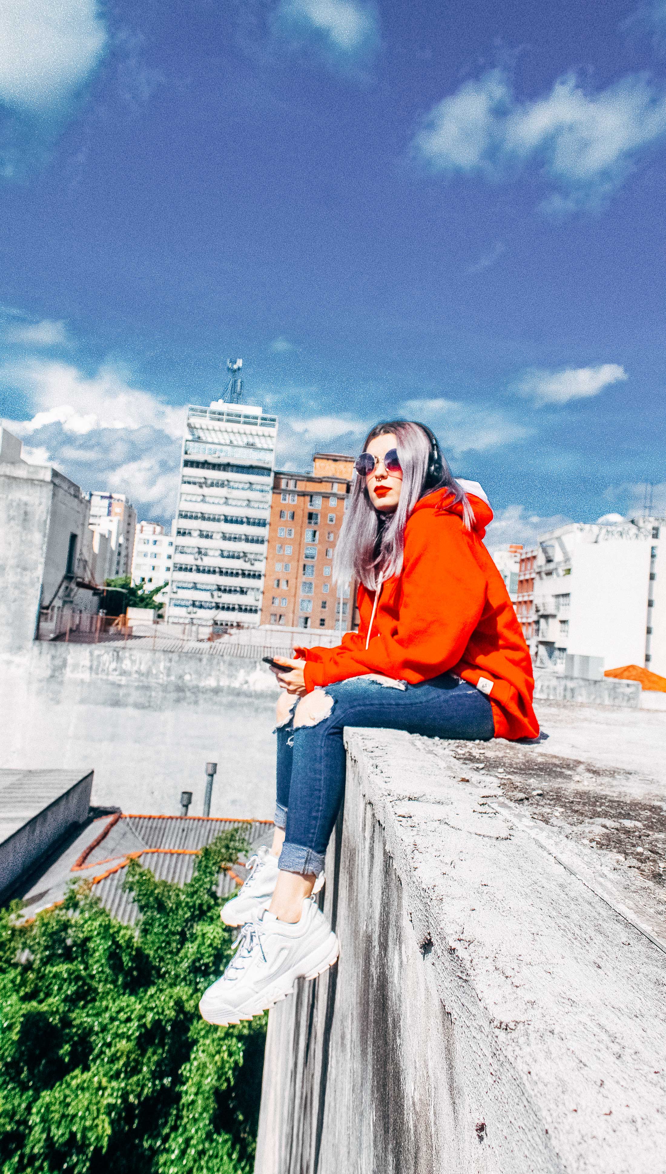 Woman sitting on a ledge facing the left, with tall buildings in the background. Below her feet are green trees. She is holding a phone or mp3 player.