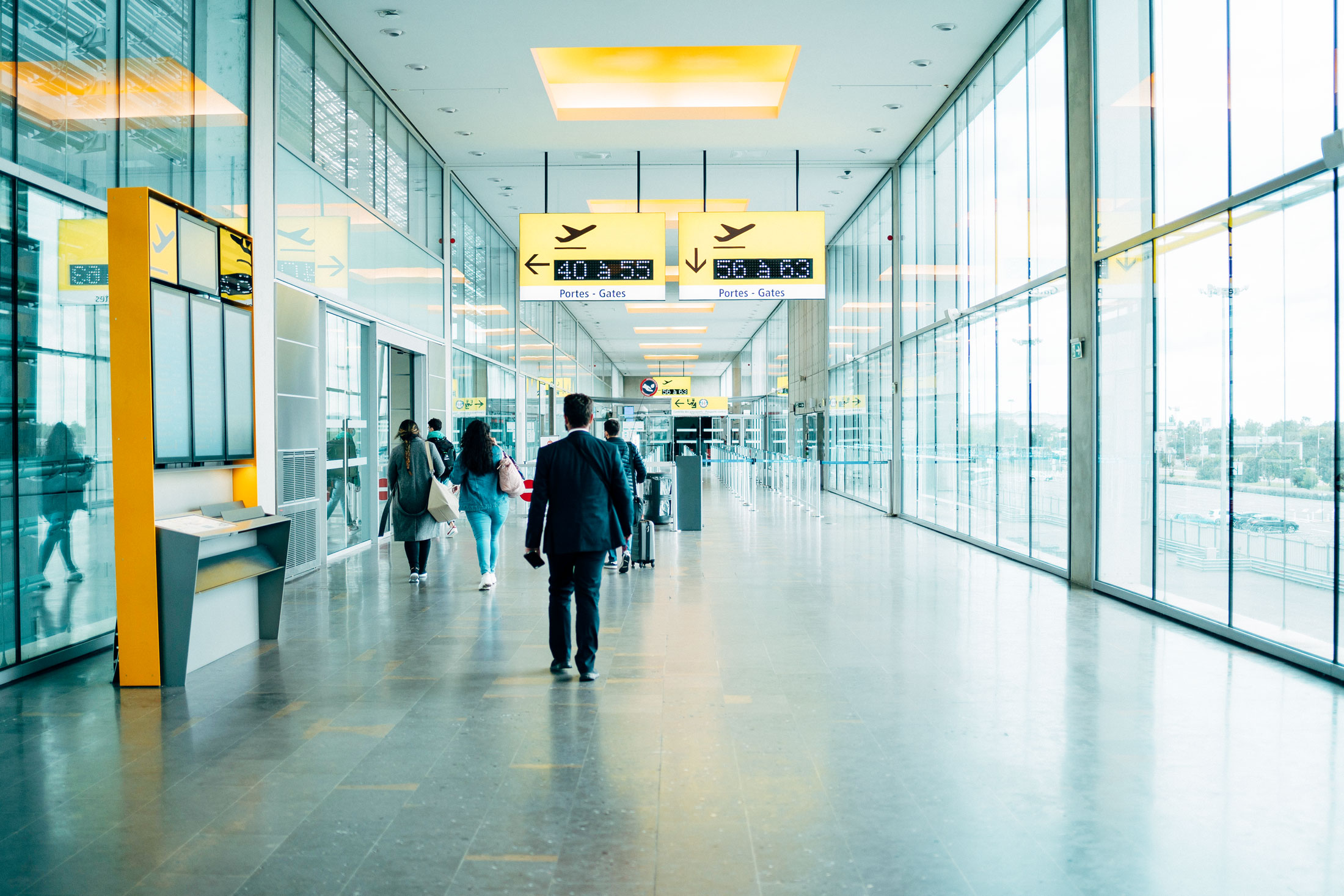 Photo showing an airport, showing glass windows on the right. Signage above a man walking down the middle. On the left are information points and a doorway.