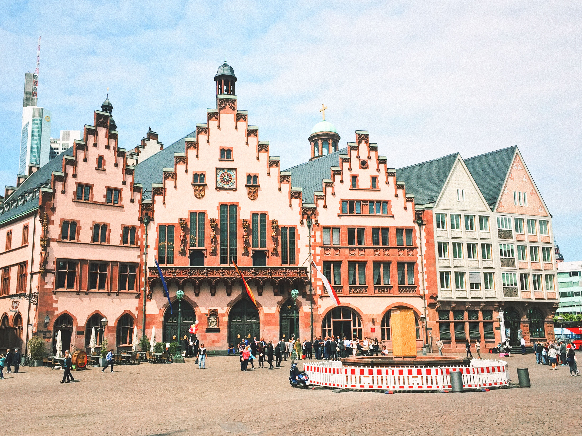 Front photo of the very traditional German Römerberg buildings. The roofs are block square constructed and the front of the buildings are light pink and dark orange. People are walking in the square in front, and there is a fountain there.