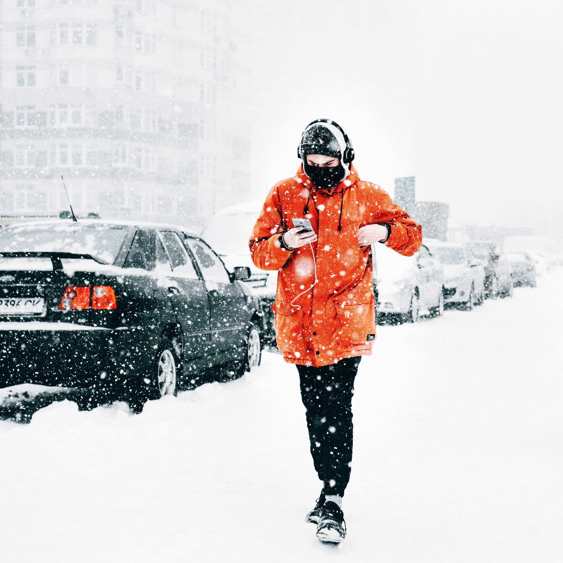 Man walking along a snow-covered pavement on the right. On the left are a line of cars that are also covered by snow. He is wearing a bright orange coat, and holding his iPhone listening to music.