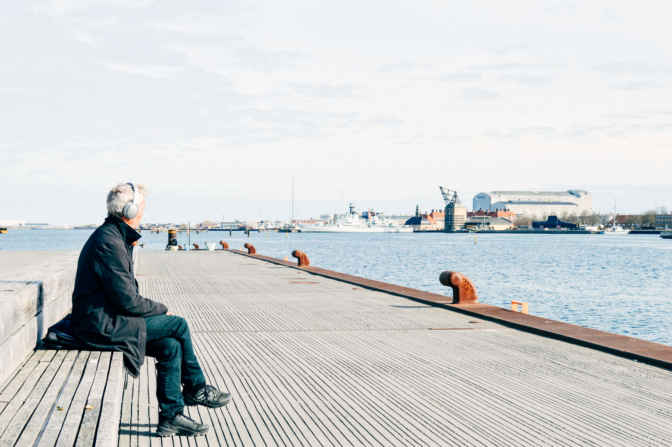 Man sitting on a ledge, on the left on a sea pier, looking to the right, that shows the sea and docklands, then factories in the far distance. The sky cover the top half.